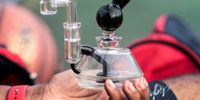 Person holding a compact glass dab rig with a quartz banger attachment and black mouthpiece, used for vaporizing cannabis concentrates. A basketball and red bag are visible in the background