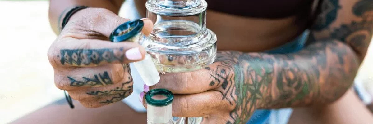 Tattooed woman outdoors holding a glass bong labeled 'Bud' while loading a bowl. The clear bong features an inline percolator and green accents, used for smoking cannabis.